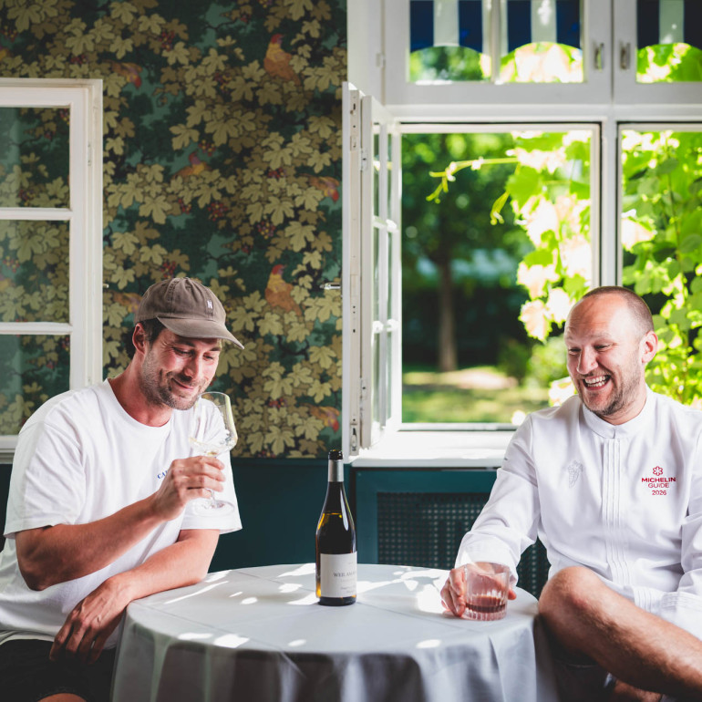 Christoph Düster und Christoph Schneider sitzen mit einer flasche Wein an einem runden Tisch.