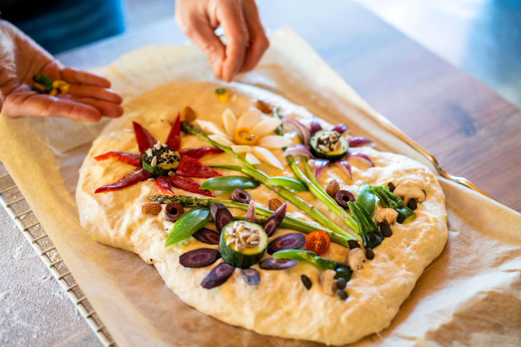 Focaccia auf Backpapier, verziert mit buntem Gemüse und Kräutern in Blumenform, während eine Person weitere Zutaten hinzufügt.