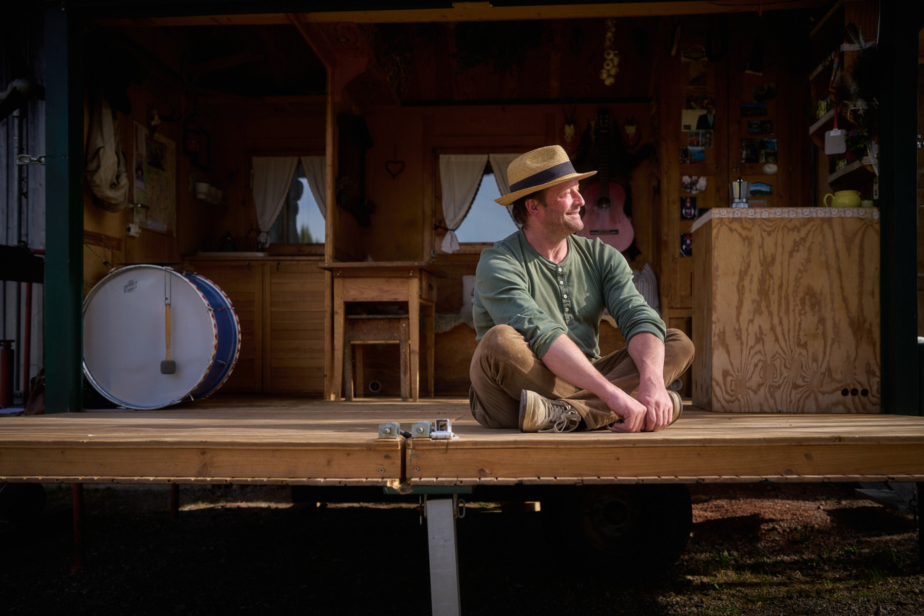 Mann mit Strohhut sitzt entspannt auf der Bühne eines nostalgischen Zirkuswagens im Schwarzwald, im Hintergrund Gitarre und große Trommel