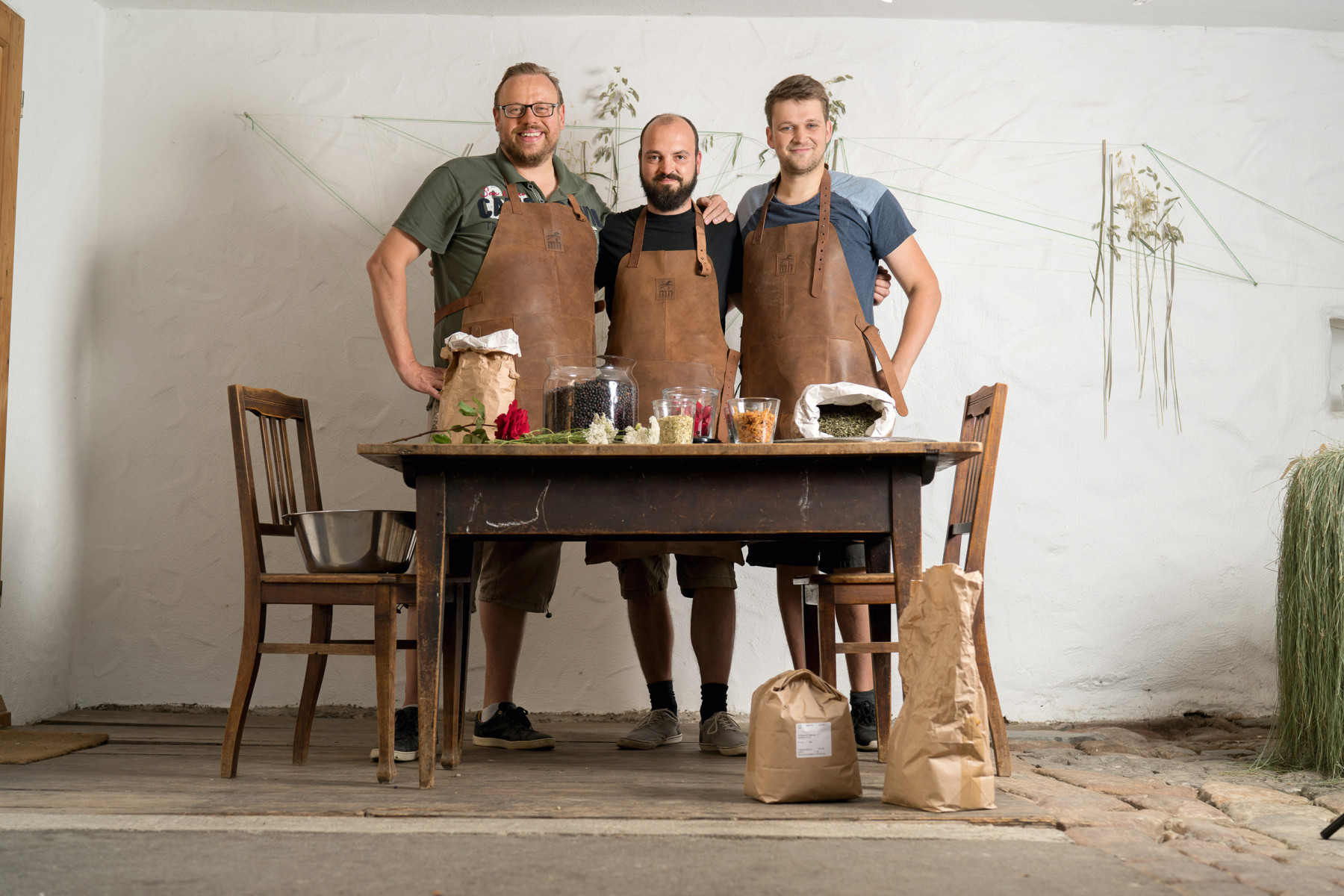 Drei Männer in Lederschürzen stehen an Holztisch mit Kräutern und Zutaten in traditioneller Werkstatt im Schwarzwald