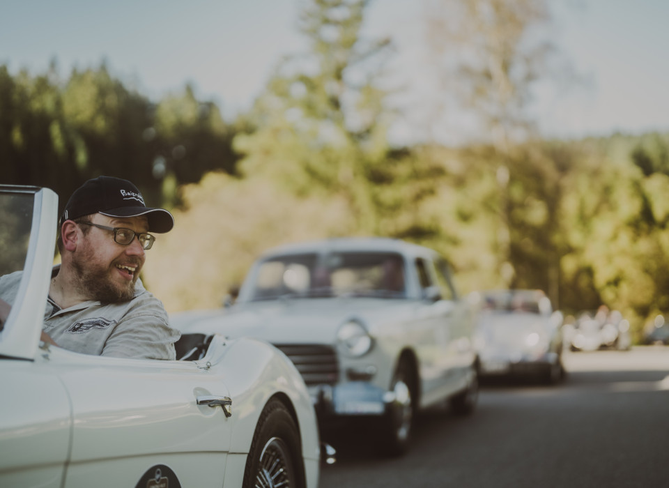 Fahrer im weißen Oldtimer-Cabrio lächelt bei Schwarzwald-Ausfahrt im Sonnenschein, mit Kolonne historischer Fahrzeuge im Hintergrund