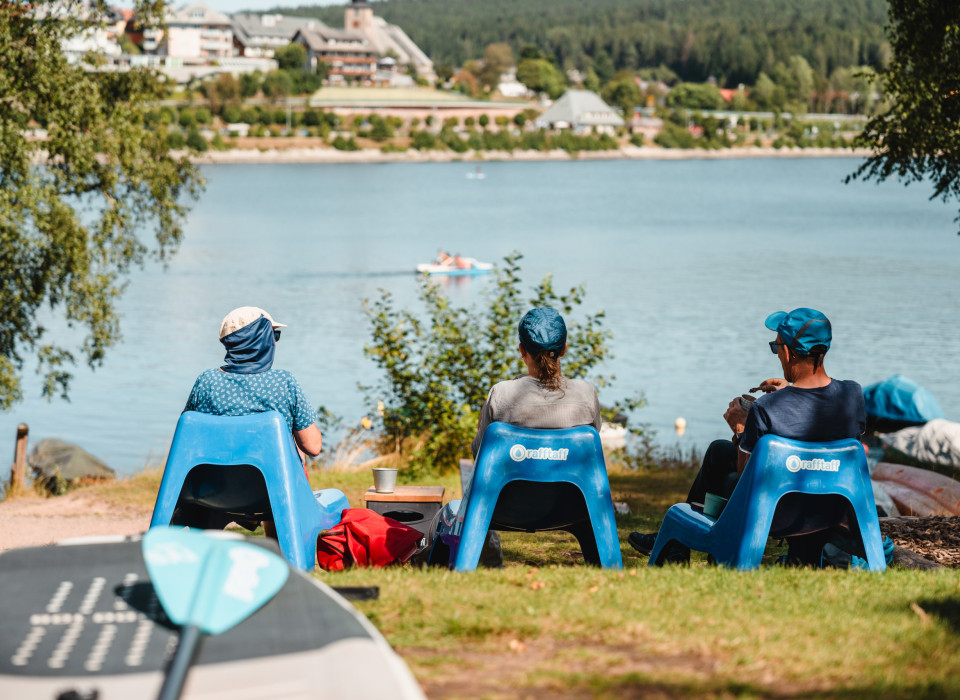 Drei Menschen entspannen am Schluchsee mit Blick auf Seepromenade und Schwarzwaldpanorama