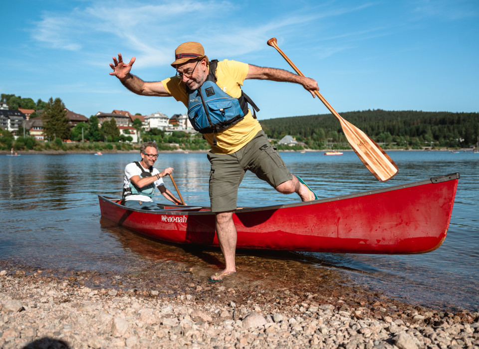 Mann springt mit Paddel aus rotem Kanu an den Kiesstrand des Schluchsees im Schwarzwald