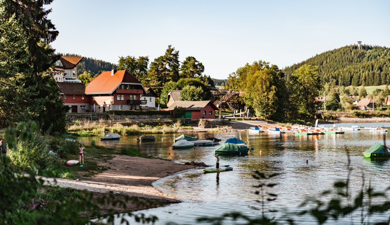 Blick auf Uferpromenade und historische Schwarzwaldhäuser am Schluchsee mit Booten und Badenden im Sommerlicht