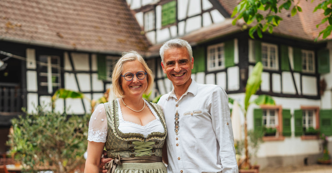 Eine Frau im Dirndl und ein Mann in Tracht stehen lächeln Arm in Arm vor einem badischen Hof.