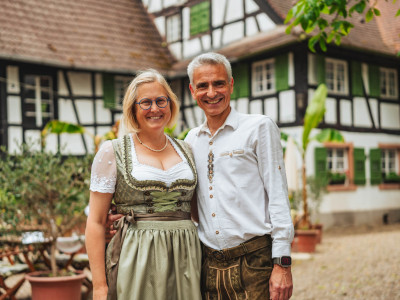 Eine Frau im Dirndl und ein Mann in Tracht stehen lächeln Arm in Arm vor einem badischen Hof.