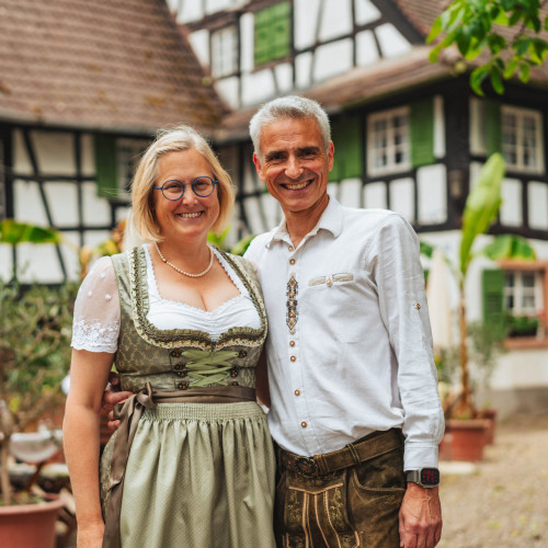 Eine Frau im Dirndl und ein Mann in Tracht stehen lächeln Arm in Arm vor einem badischen Hof.
