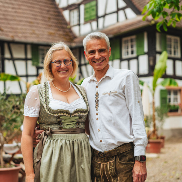 Eine Frau im Dirndl und ein Mann in Tracht stehen lächeln Arm in Arm vor einem badischen Hof.