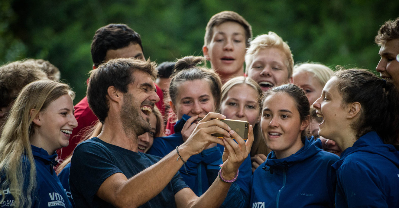 Eine Gruppe junger, vergnügter Menschen sieht sich gemeinsam etwas auf einem Smartphone an. Sie befinden sich in der Wildnis. 