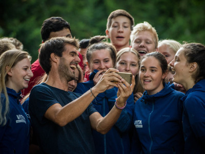 Eine Gruppe junger, vergnügter Menschen sieht sich gemeinsam etwas auf einem Smartphone an. Sie befinden sich in der Wildnis. 
