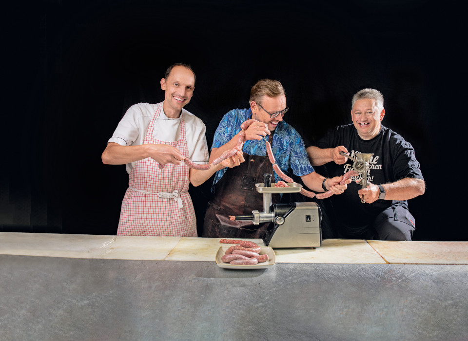 Drei Männer lachen beim Wurstmachen an der Fleischmaschine – handgemachte Heimatwurst mit Spaßfaktor
