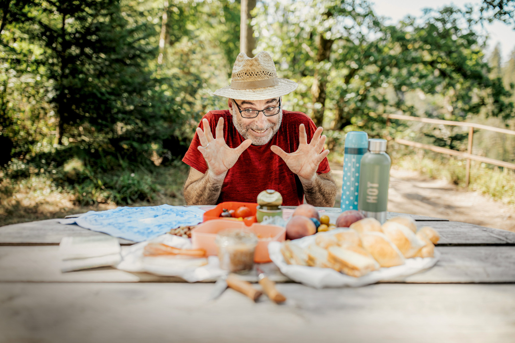 Älterer Mann mit Strohhut und rotem Shirt am gedeckten Picknicktisch im Wald, scherzhaft mit erhobenen Händen