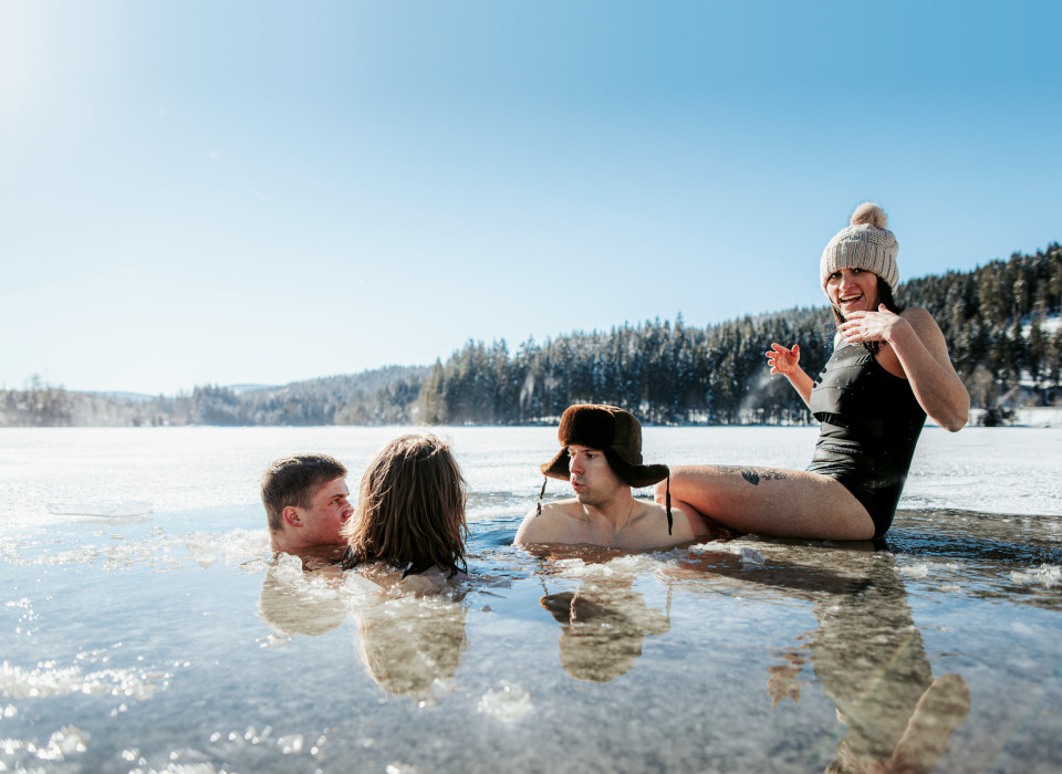 Vier Menschen beim Eisbaden in gefrorenem Schwarzwaldsee, lachend im Sonnenlicht mit Mütze und Badeanzug