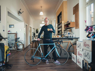 Fröhlicher Mann steht in seinem Café, vor sich ein pepsiblauen Rennrad. In der Hand hält er eine Espressotasse