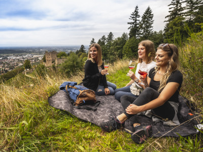 Drei Frauen machen eine Wanderpause und trinken ein Glas Wein. Im Hintergrund ist die Burgruine Schauenburg