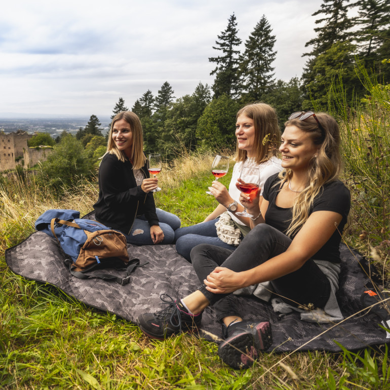 Drei Frauen machen eine Wanderpause und trinken ein Glas Wein. Im Hintergrund ist die Burgruine Schauenburg