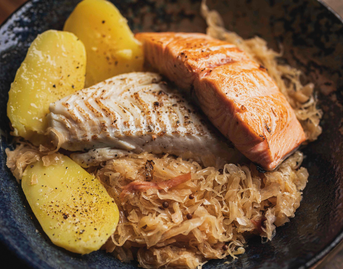 Sauerkraut mit Fisch und Kartoffeln auf einem grauen Teller, serviert auf einem Holzbrett.