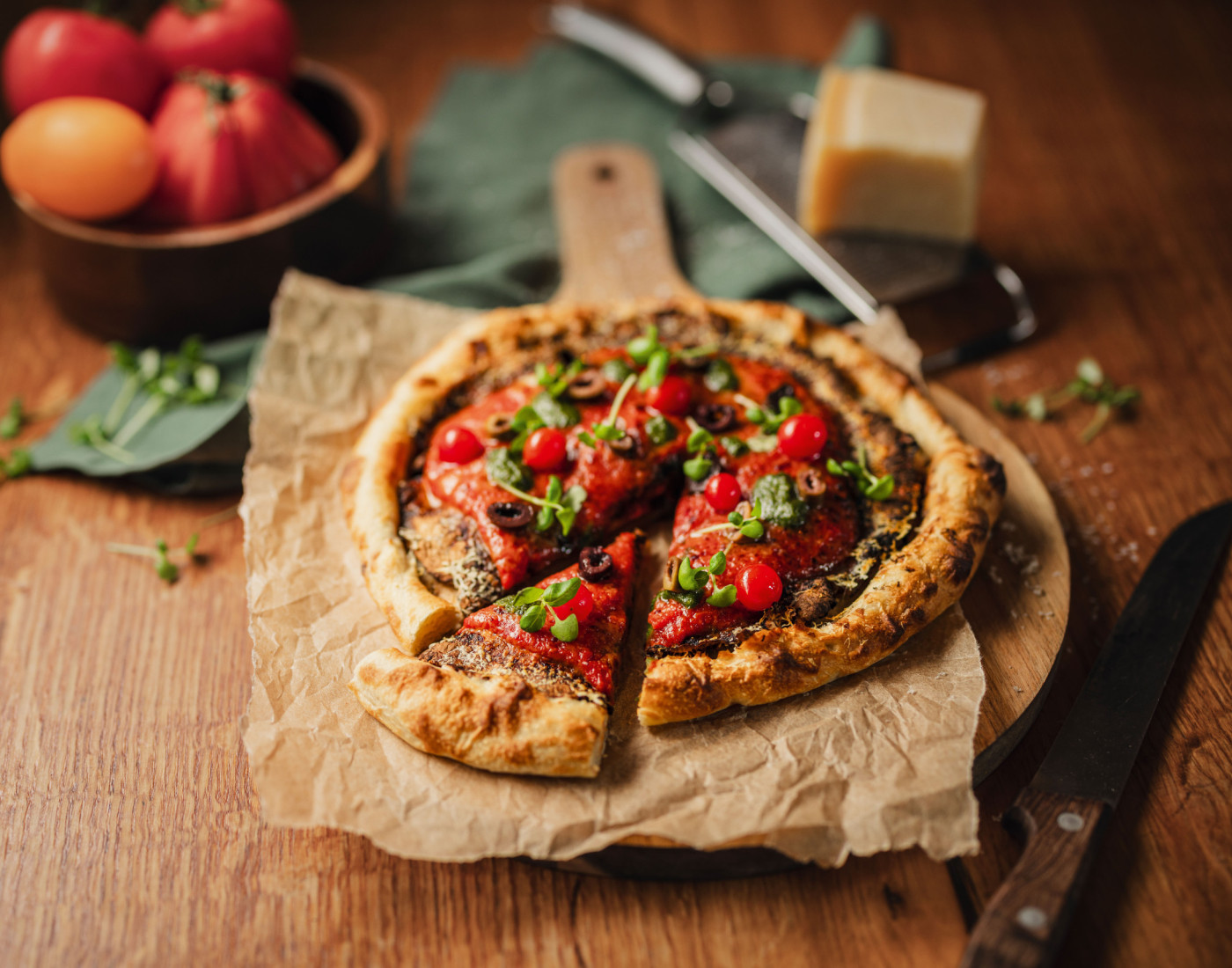 Pizza Parasol mit Tomatenkompott, serviert auf Holzbrett mit Backpapier, im Hintergrund Käse und Gemüse