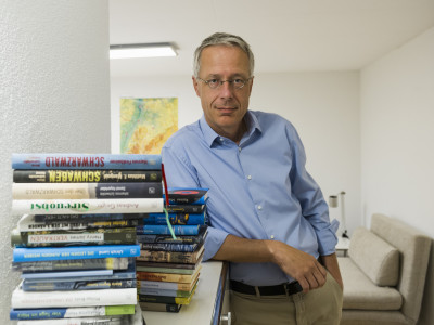 Der Freiburger Verleger steht hinter einem Stapel Bücher und schaut frontal in die Kamera