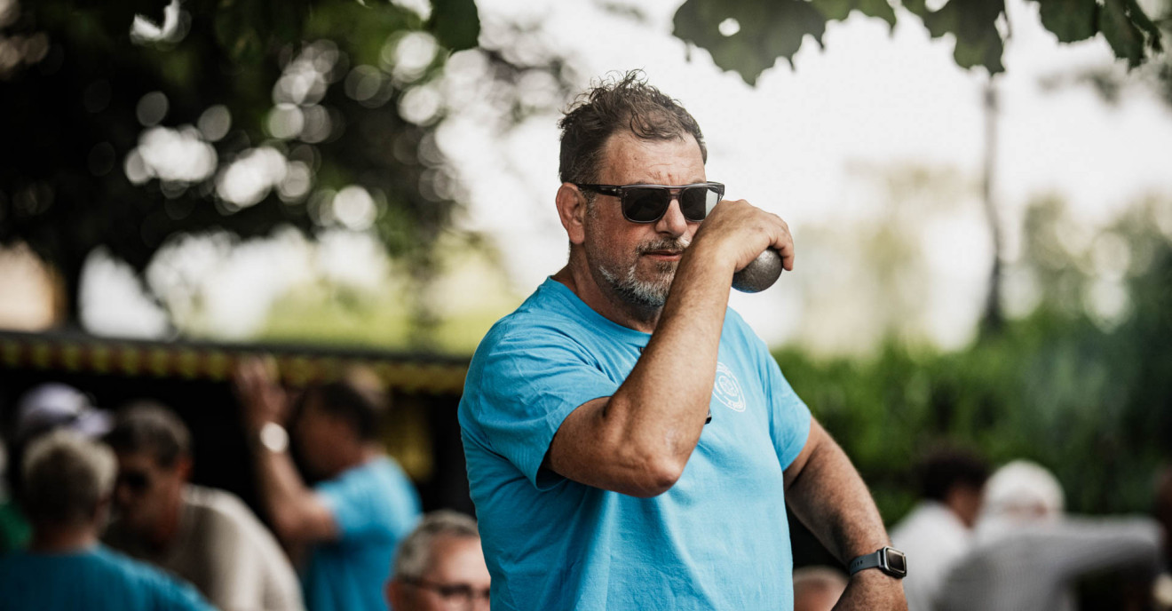Ein Mann in einem hellblauen T-Shirt holt aus, um eine Boule-Kugel zu werfen.