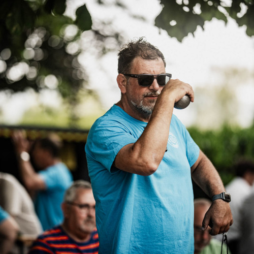 Ein Mann in einem hellblauen T-Shirt holt aus, um eine Boule-Kugel zu werfen.