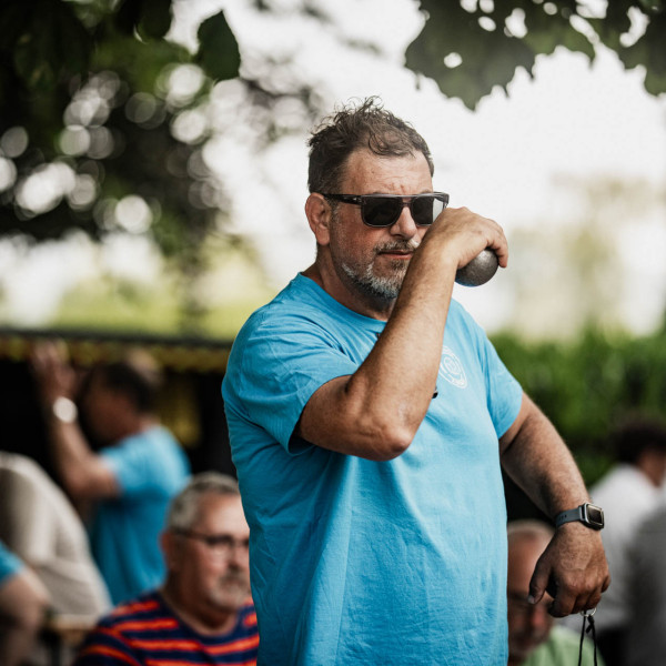 Ein Mann in einem hellblauen T-Shirt holt aus, um eine Boule-Kugel zu werfen.