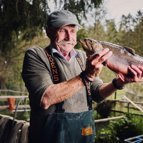 Ein Forellenzüchter hält einen Fisch in die Höhe und lächelt.