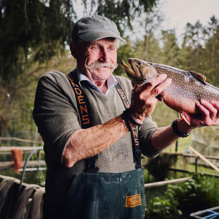 Ein Forellenzüchter hält einen Fisch in die Höhe und lächelt.
