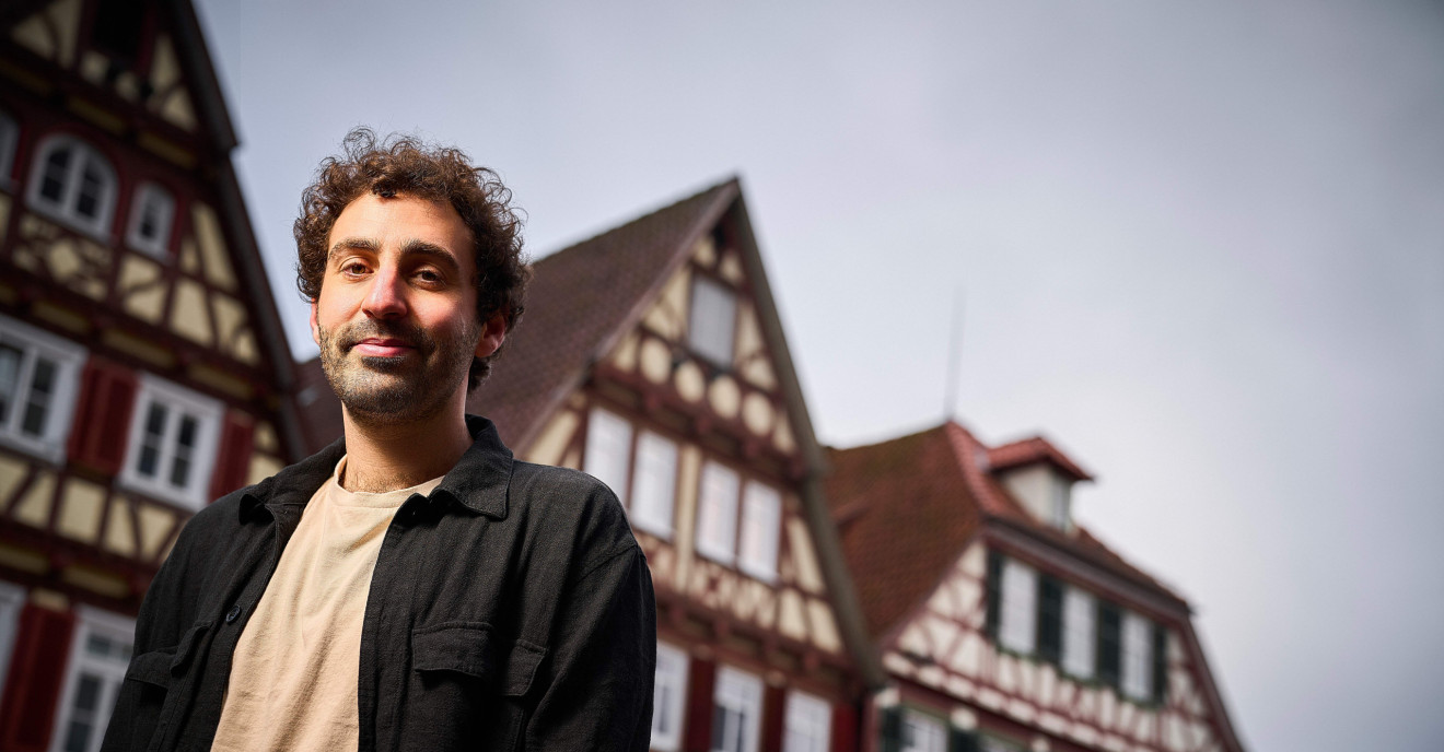 Ein junger Mann mit dunklen, kurzen Locken steht vor typisch süddeutschen Fachwerkhäusern.