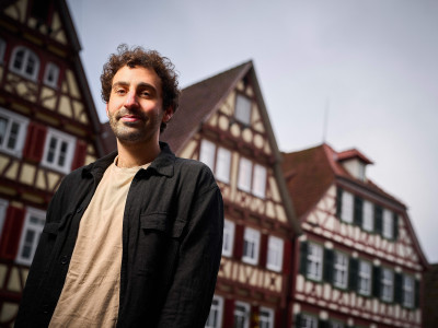 Ein junger Mann mit dunklen, kurzen Locken steht vor typisch süddeutschen Fachwerkhäusern.
