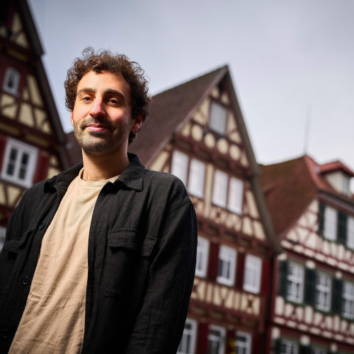Ein junger Mann mit dunklen, kurzen Locken steht vor typisch süddeutschen Fachwerkhäusern.