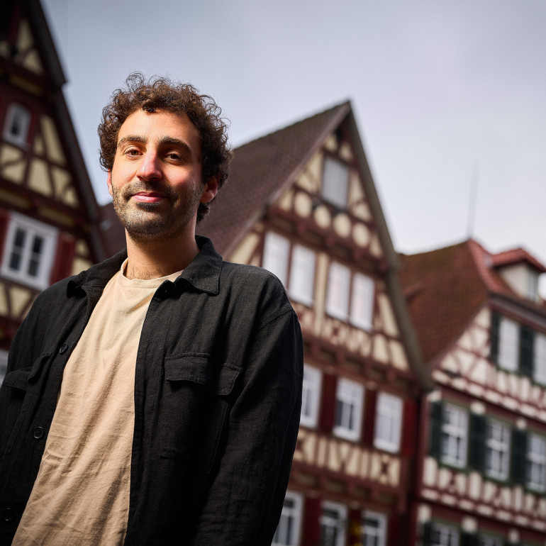 Ein junger Mann mit dunklen, kurzen Locken steht vor typisch süddeutschen Fachwerkhäusern.
