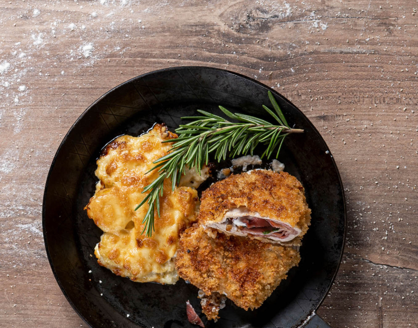 Ein Cordon Bleu, Kartoffelgratin und Rosmarin wurden auf einer dunklen Eisenpfanne angerichtet.