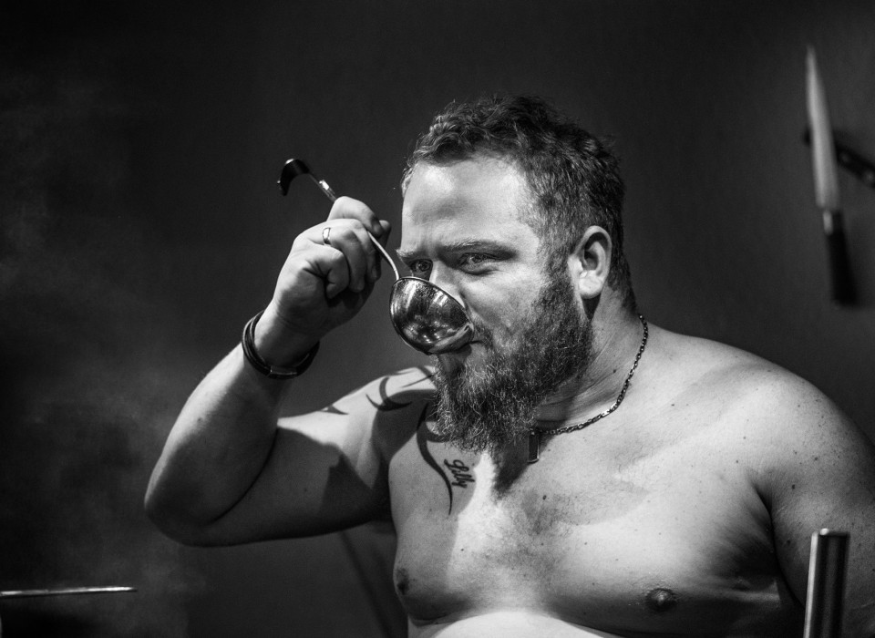 Oben ohne Koch mit Suppenkelle beim Probieren am Herd – künstlerisches Schwarz-Weiß-Porträt aus der Serie „The Naked Cook“